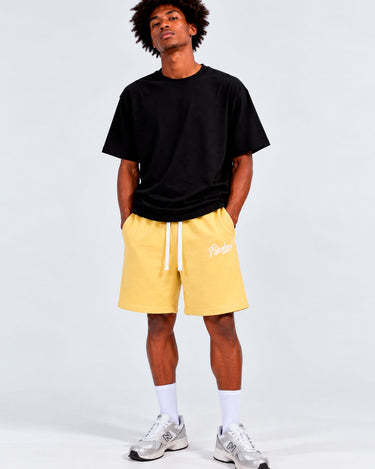 Pantalones cortos amarillos con cordón blanco, camiseta negra, modelo de pie, fondo gris claro.