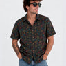 Camisa de hombre negra con estampado colorido, botones frontales y mangas cortas, combinada con gafas de sol.