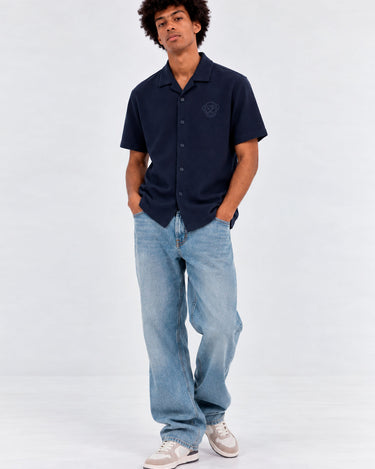 Camisa unisex de color navy con botones y logo en el pecho, combinada con jeans claros y zapatillas. Estilo casual.