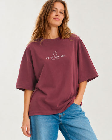 Camiseta boxy unisex color púrpura con texto blanco "THE TRIP IS THE TRUTH" en el pecho, modelo femenino sonriente