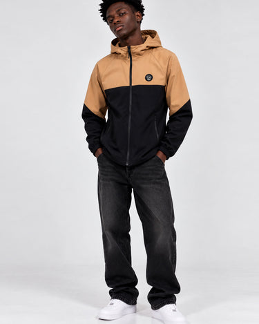 Cortavientos impermeable en negro y caramelo, con capucha y logo visible en el pecho, modelo de pie.