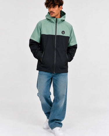 Cortavientos impermeable en negro y verde, con capucha y logo visible en el pecho, modelo masculino con jeans claros.