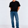 Pantalón vaquero azul claro en hombre, camiseta negra, fondo blanco, zapatos blancos.