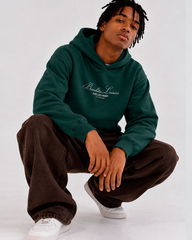 Sudadera unisex verde oscuro con bordado 'Bendita Locura' y logo de Loco Monky, modelo sentado con pantalones oscuros.