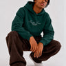 Sudadera unisex verde oscuro con bordado 'Bendita Locura' y logo de Loco Monky, modelo sentado con pantalones oscuros.