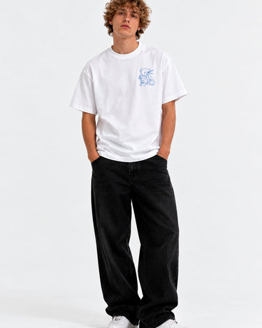Camiseta blanca unisex con diseño azul en el pecho, modelo de pie con pantalones negros y zapatillas blancas.