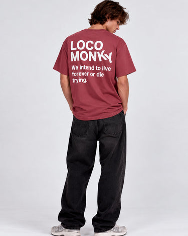 Camiseta de color burdeos con texto blanco en la espalda, hombre de pie con pantalones negros.