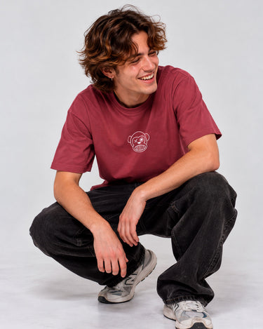 Camiseta de color burdeos con un diseño de calavera en el pecho, modelo masculino sonriente en pantalones oscuros.