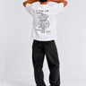 Camiseta blanca con diseño de mono y texto en la parte trasera, pantalones oscuros y fondo neutro.