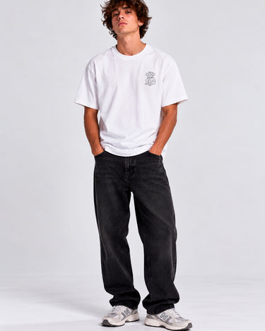 Camiseta blanca con diseño en el pecho, modelo masculino de pie, pantalones oscuros y zapatillas deportivas.