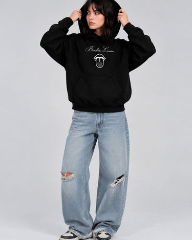 Sudadera negra unisex con diseño en el pecho, combinada con jeans claros y zapatillas deportivas.
