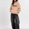 Sudadera unisex color beige con capucha y estampado en negro, combinada con pantalones oscuros y zapatillas blancas.