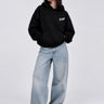 Sudadera oversize negra con capucha, logo blanco en el pecho, combinada con jeans claros y zapatillas blancas.