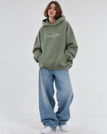 Sudadera boxy unisex en color oliva con capucha y texto blanco en el pecho, combinada con jeans claros.