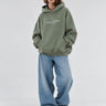 Sudadera boxy unisex en color oliva con capucha y texto blanco en el pecho, combinada con jeans claros.