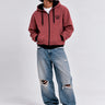 Sudadera con cremallera color vino, con capucha y logo en el pecho, combinada con jeans desgastados.