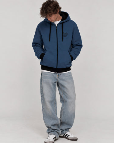 Sudadera con cremallera azul profundo, capucha, detalles en negro, modelo masculino con jeans claros.