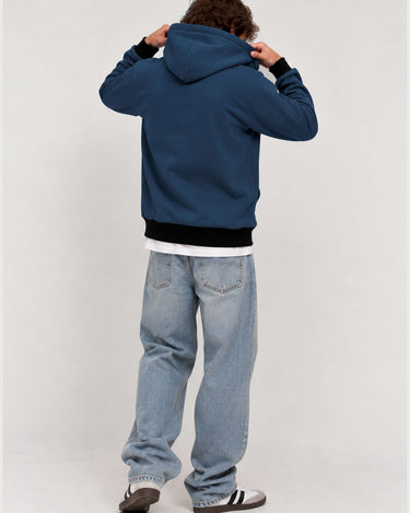 Sudadera con capucha de color azul profundo, vista trasera, con pantalones de mezclilla claros y zapatos deportivos.