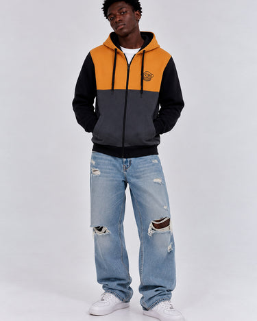 Sudadera con cremallera en mostaza, negro y gris oscuro, con capucha y logo en el pecho, modelo masculino con jeans rotos.