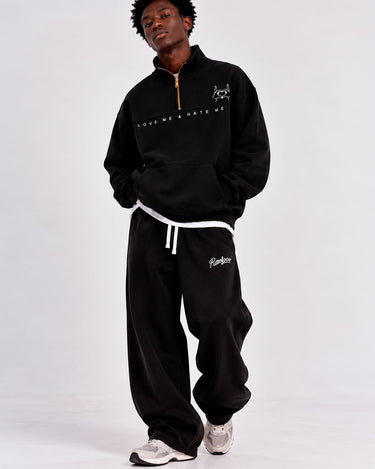 Sudadera oversize negra con cremallera y texto 'LOVE ME & HATE ME', pantalones negros, modelo de pie.
