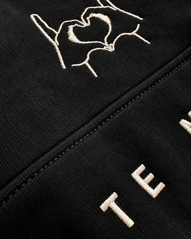 Sudadera negra con diseño bordado de manos formando un corazón y texto en la parte inferior.