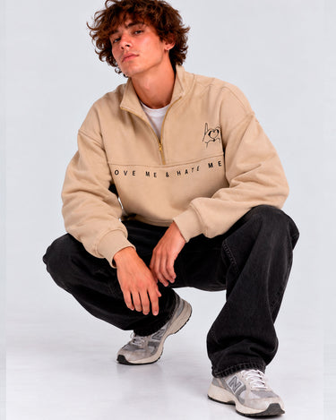 Sudadera oversize beige con logo bordado 'LOVE ME & HATE ME' en el pecho, modelo sentado, jeans negros.