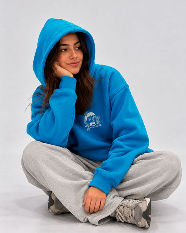 Sudadera azul con capucha, estampado frontal de Loco Monky, modelo sentado con pantalones grises.