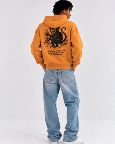 Sudadera unisex mostaza con diseño de mono y texto en la parte posterior, jeans claros y fondo neutro.