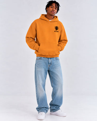 Sudadera unisex color mostaza con capucha y logo negro en el pecho, combinada con jeans claros.