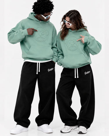Pantalones oversize negros con cordones, combinados con sudaderas verdes, dos modelos posando juntos.