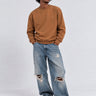 Sudadera color caramelo con logo en el pecho, jeans desgastados y zapatillas blancas, fondo gris claro.