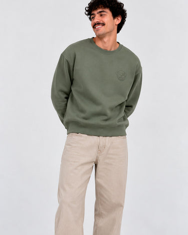 Sudadera de color oliva con logo en el pecho, modelo masculino con pantalones claros y fondo neutro.