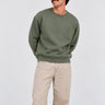 Sudadera de color oliva con logo en el pecho, modelo masculino con pantalones claros y fondo neutro.