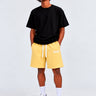 Pantalones cortos amarillos con cordón blanco, camiseta negra, modelo de pie, fondo gris claro.