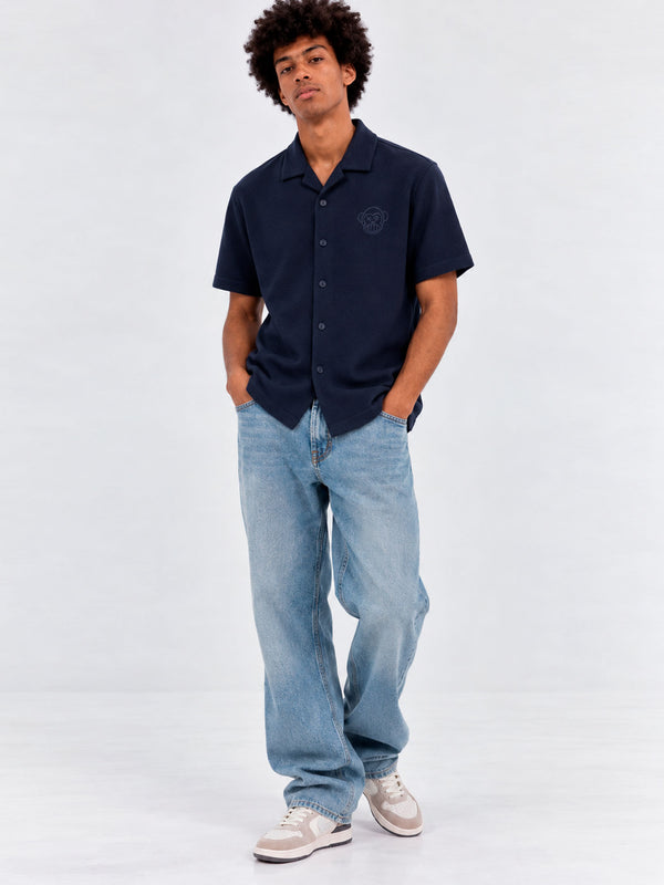 Camisa unisex de color navy con botones y logo en el pecho, combinada con jeans claros y zapatillas. Estilo casual.