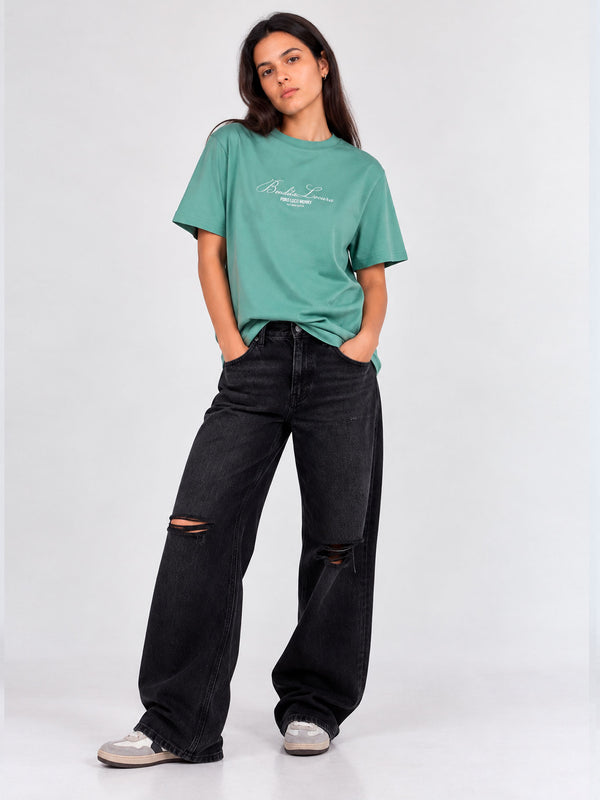 Camiseta verde con bordado en el pecho, combinada con pantalones negros rotos y calzado claro.