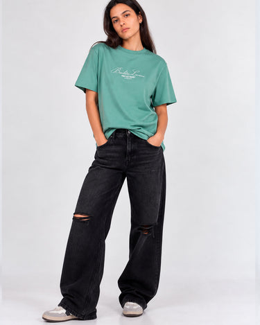 Camiseta verde con bordado en el pecho, combinada con pantalones negros rotos y calzado claro.