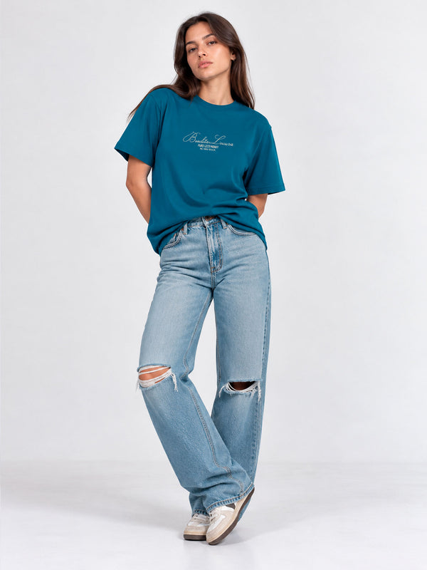 Camiseta azul con bordado en el pecho, mujer de pie con jeans rotos y zapatillas, fondo claro.