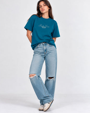 Camiseta azul con bordado en el pecho, mujer de pie con jeans rotos y zapatillas, fondo claro.