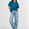 Camiseta azul con bordado en el pecho, mujer de pie con jeans rotos y zapatillas, fondo claro.