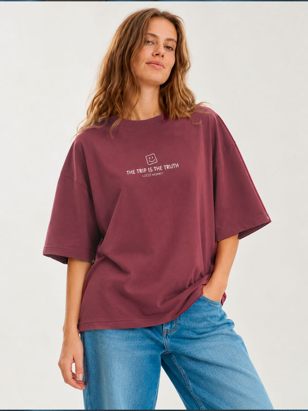Camiseta boxy unisex color púrpura con texto blanco "THE TRIP IS THE TRUTH" en el pecho, modelo femenino sonriente
