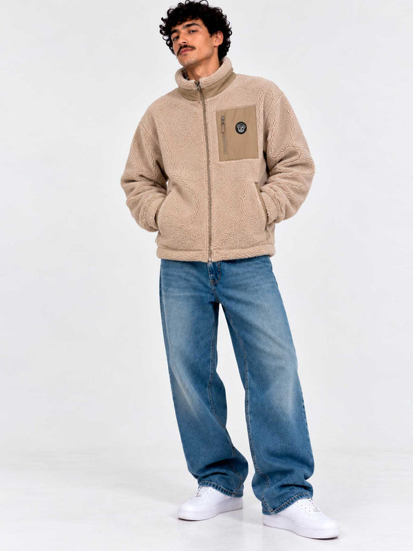 Chaqueta Sherpa unisex color beige con cremallera y bolsillo frontal, modelo con jeans y zapatillas blancas.