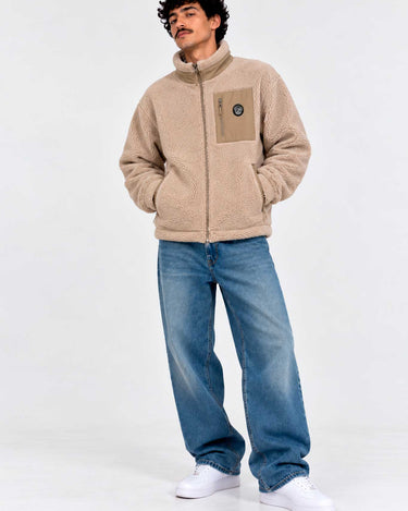 Chaqueta Sherpa unisex color beige con cremallera y bolsillo frontal, modelo con jeans y zapatillas blancas.