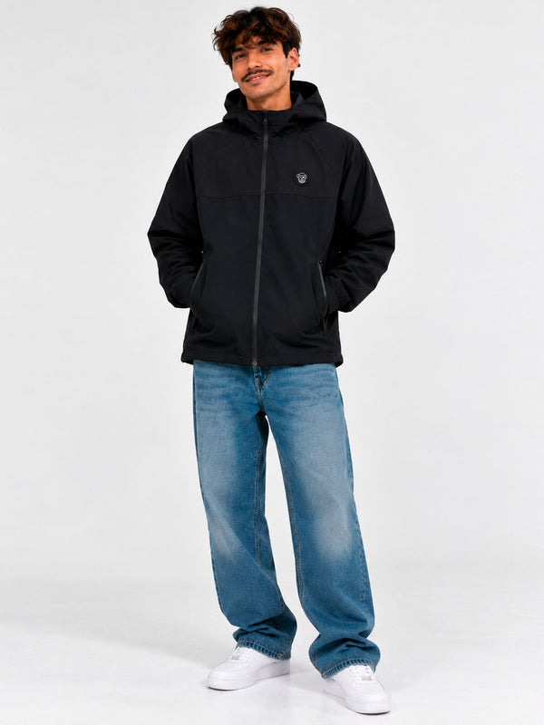 Cortavientos impermeable negro con capucha, logo en el pecho, pantalones de mezclilla claros, fondo neutro.