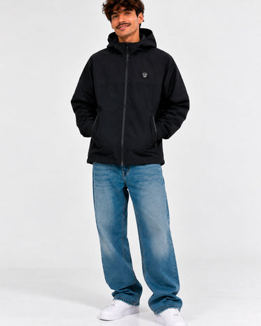 Cortavientos impermeable negro con capucha, logo en el pecho, pantalones de mezclilla claros, fondo neutro.