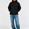 Cortavientos impermeable negro con capucha, logo en el pecho, pantalones de mezclilla claros, fondo neutro.