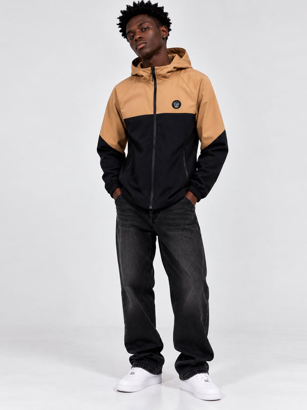 Cortavientos impermeable en negro y caramelo, con capucha y logo visible en el pecho, modelo de pie.