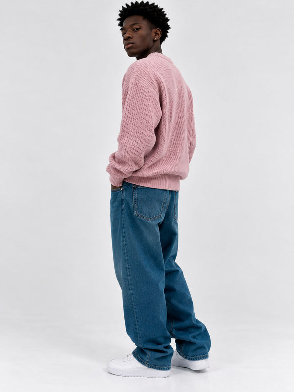Jersey de hombre color rosa, tejido grueso, con cuello redondo, combinado con jeans azules y zapatillas blancas.