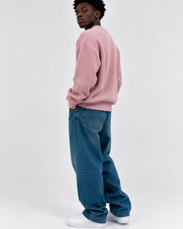 Jersey de hombre color rosa, tejido grueso, con cuello redondo, combinado con jeans azules y zapatillas blancas.