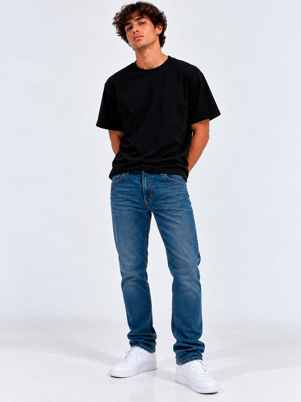 Pantalón vaquero azul claro en hombre, camiseta negra, fondo blanco, zapatos blancos.
